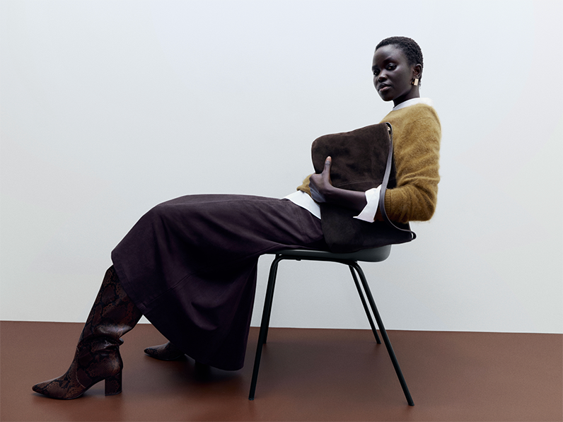 Woman wearing mustard jumper over white shirt with long brown skirt and tall brown boots, holding large clutch bag and sitting on chair