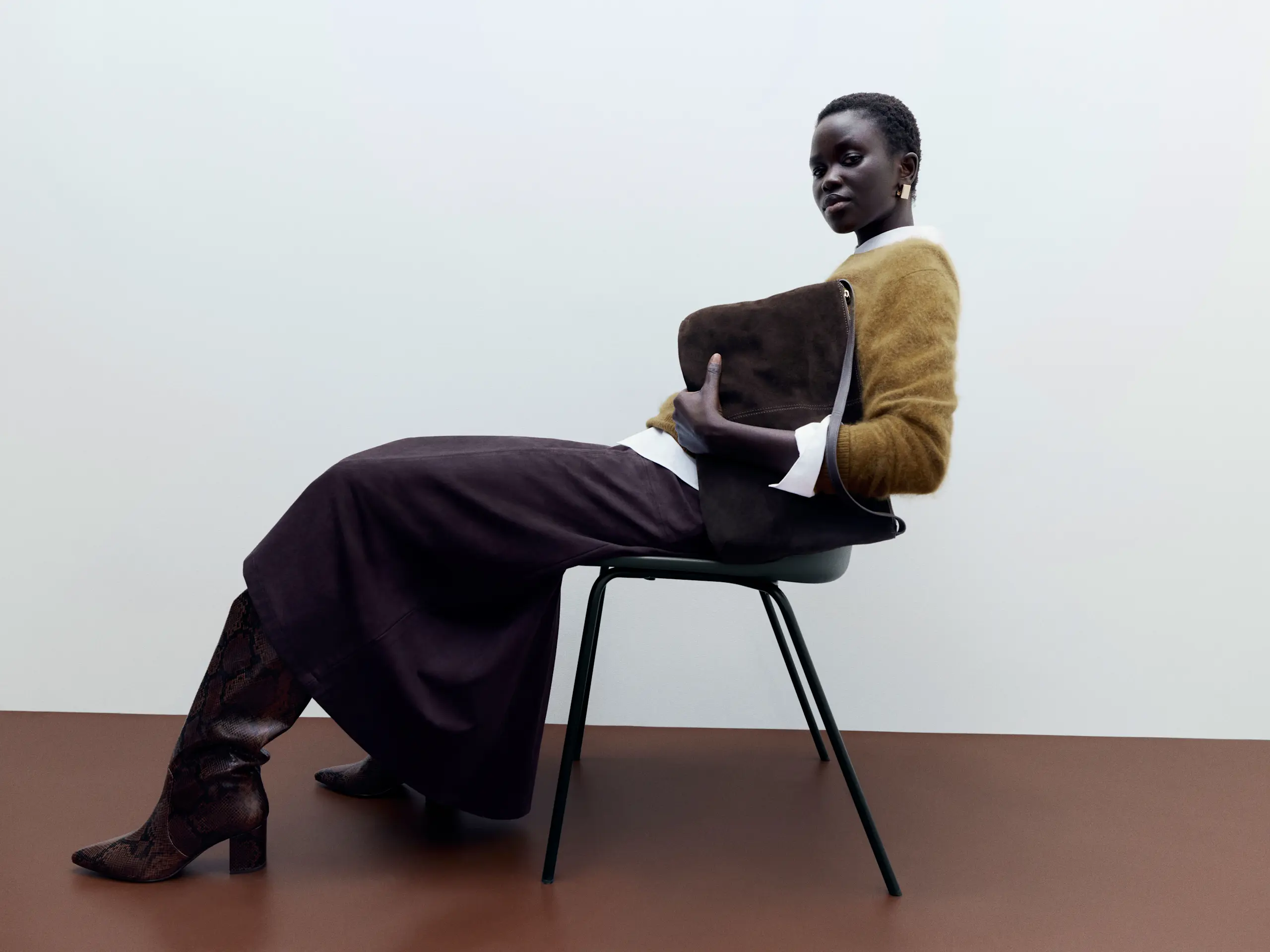 Woman wearing mustard jumper over white shirt with long brown skirt and tall brown boots, holding large clutch bag and sitting on chair