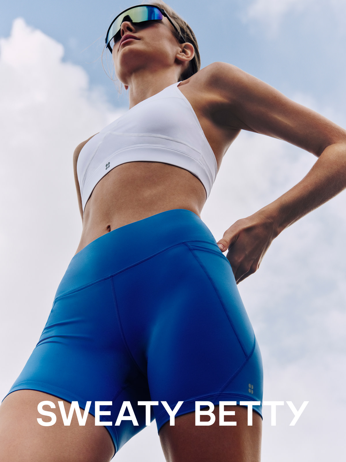 Woman wearing white sports bra, blue shorts and sunglasses by Sweaty Betty