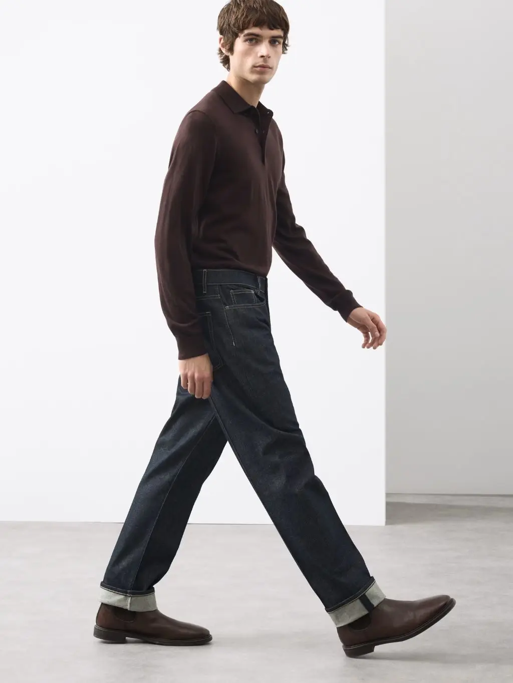 A man wears a brown jumper, dark jeans and brown chelsea boots