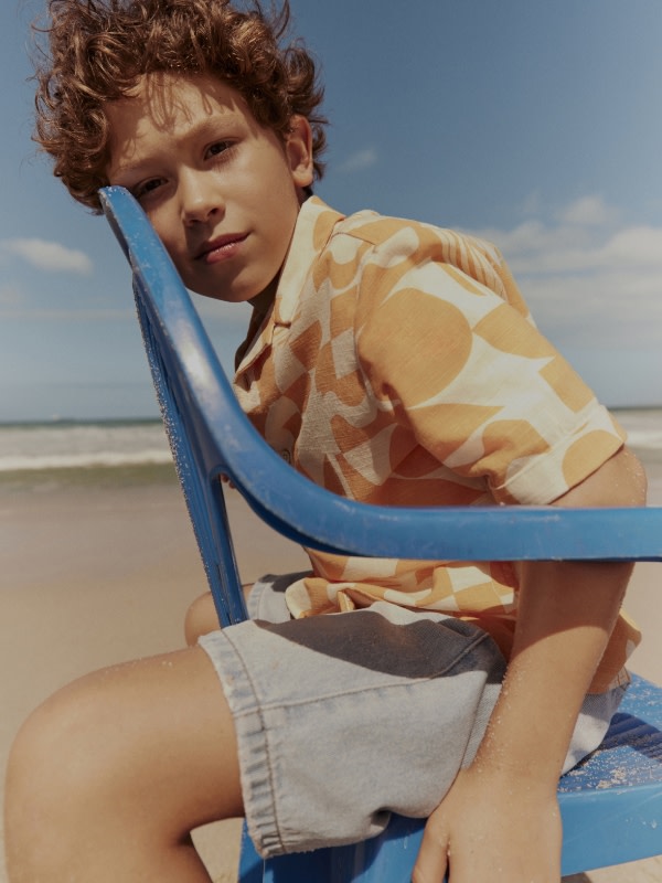 Boy wearing an orange and white patterned short-sleeved shirt and shorts. Shop boys’ shorts
