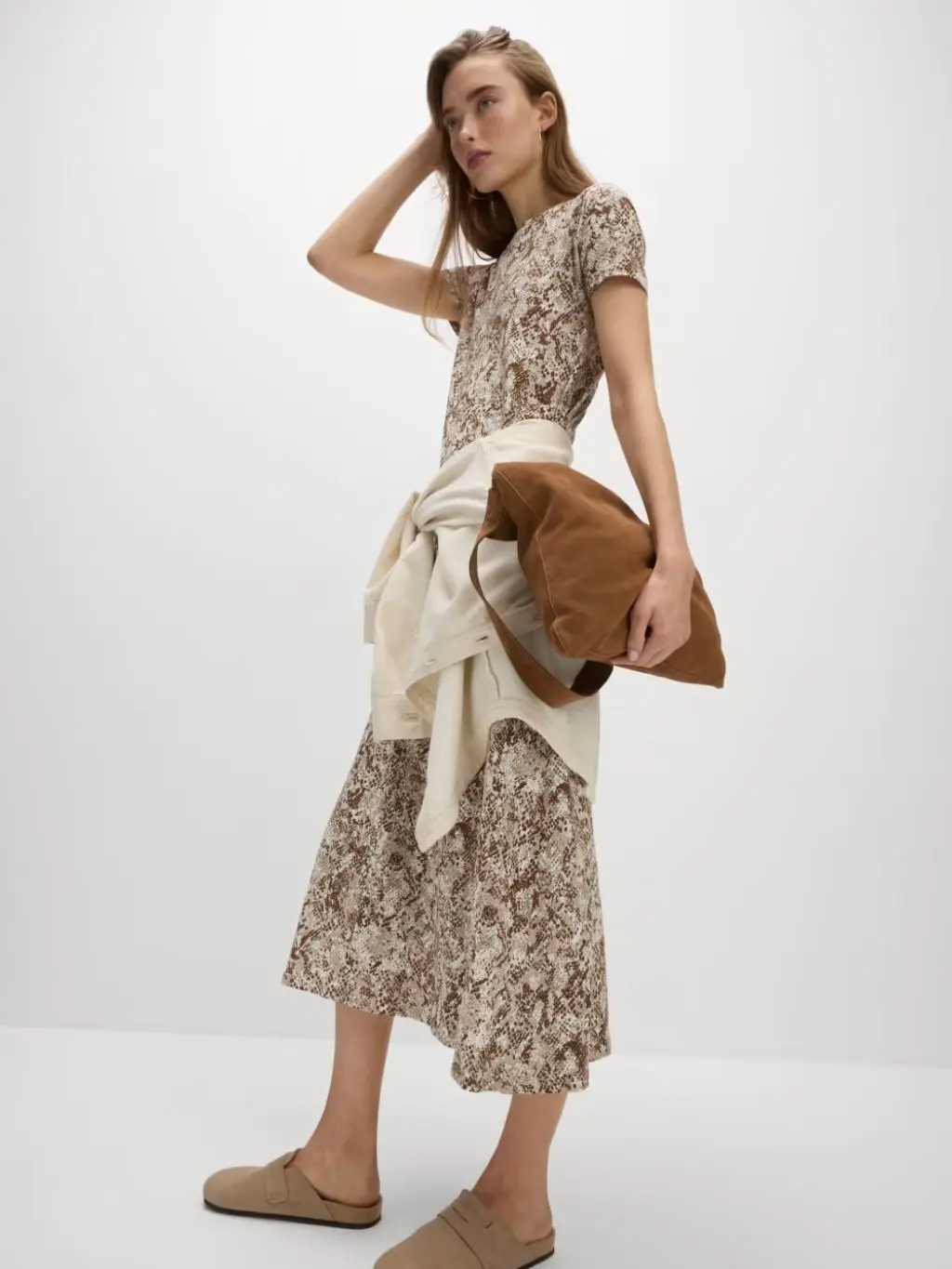 A woman wears a patterned midi T-shirt dress with a jacket tied around her waist, carrying a brown handbag