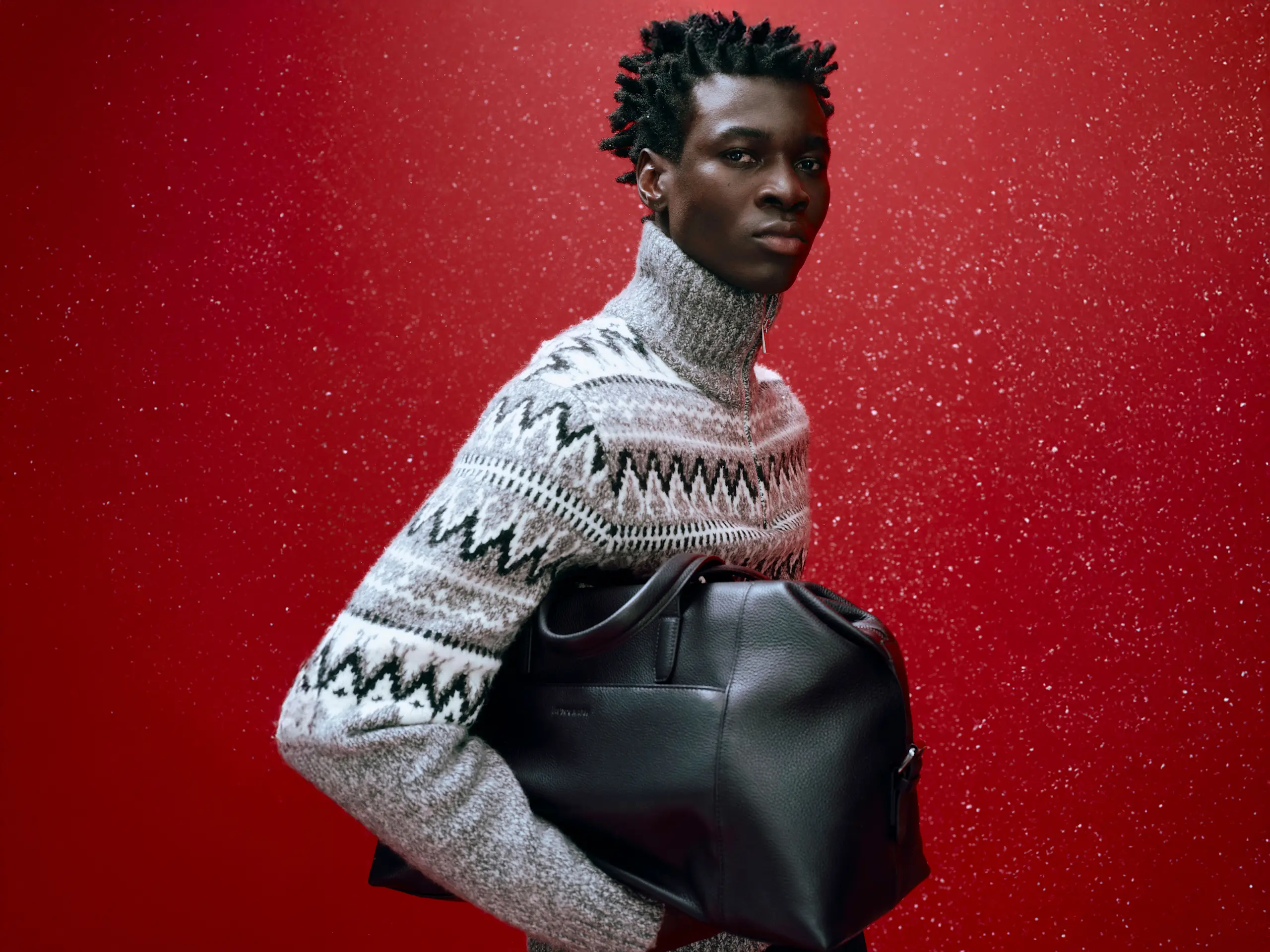 Man wearing a patterned quart-zip jumper with a black leather bag against a sparkly red backdrop