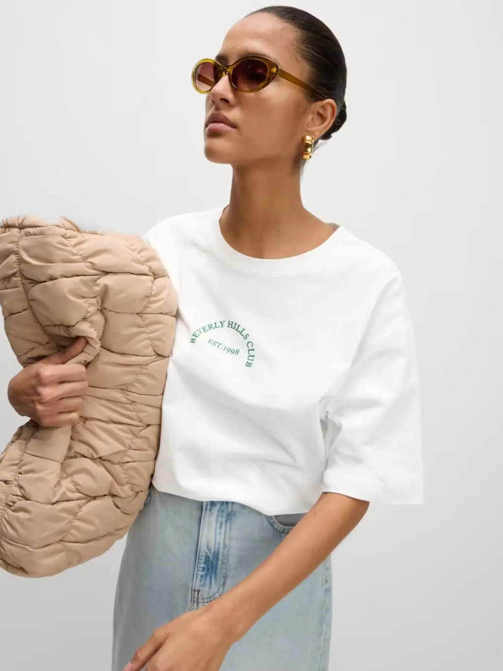 A woman wears sunglasses, a white oversized T-shirt, denim skirt and carries a beige handbag