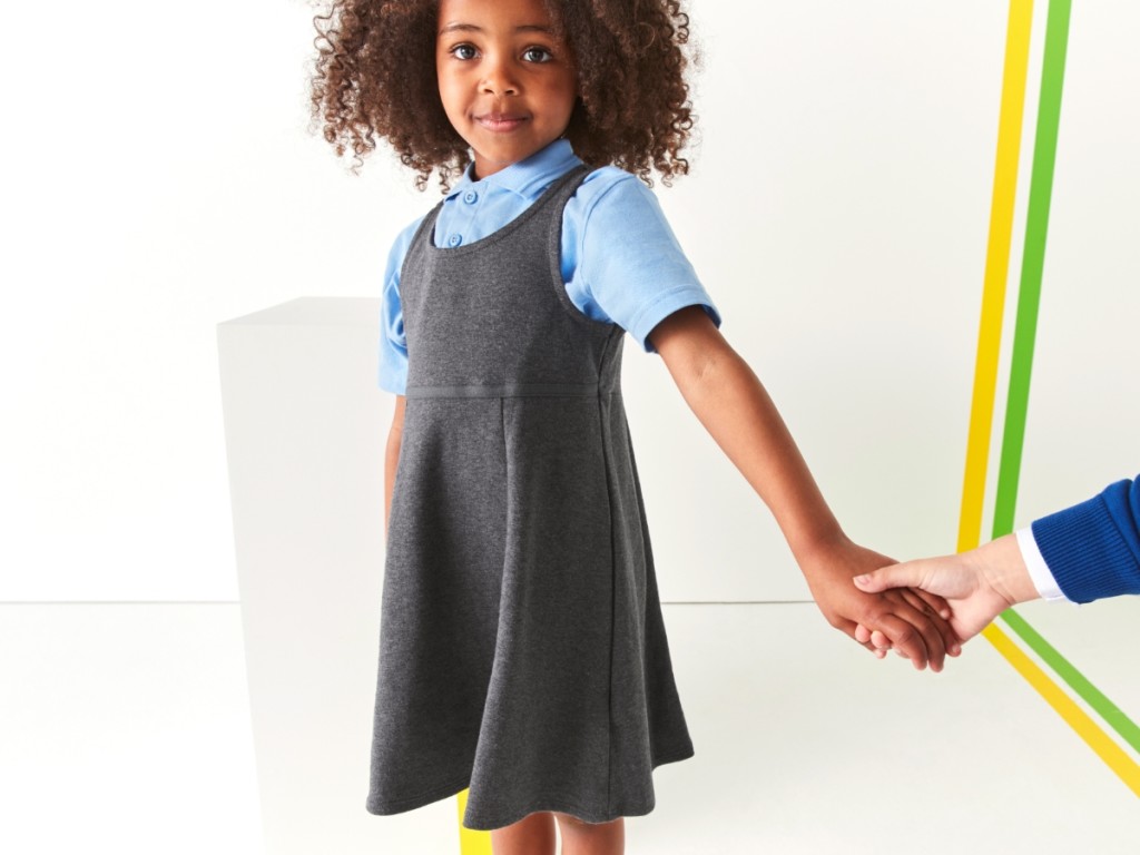 Girl wearing a blue short-sleeved shirt and grey pinafore. Shop girls’ school uniform