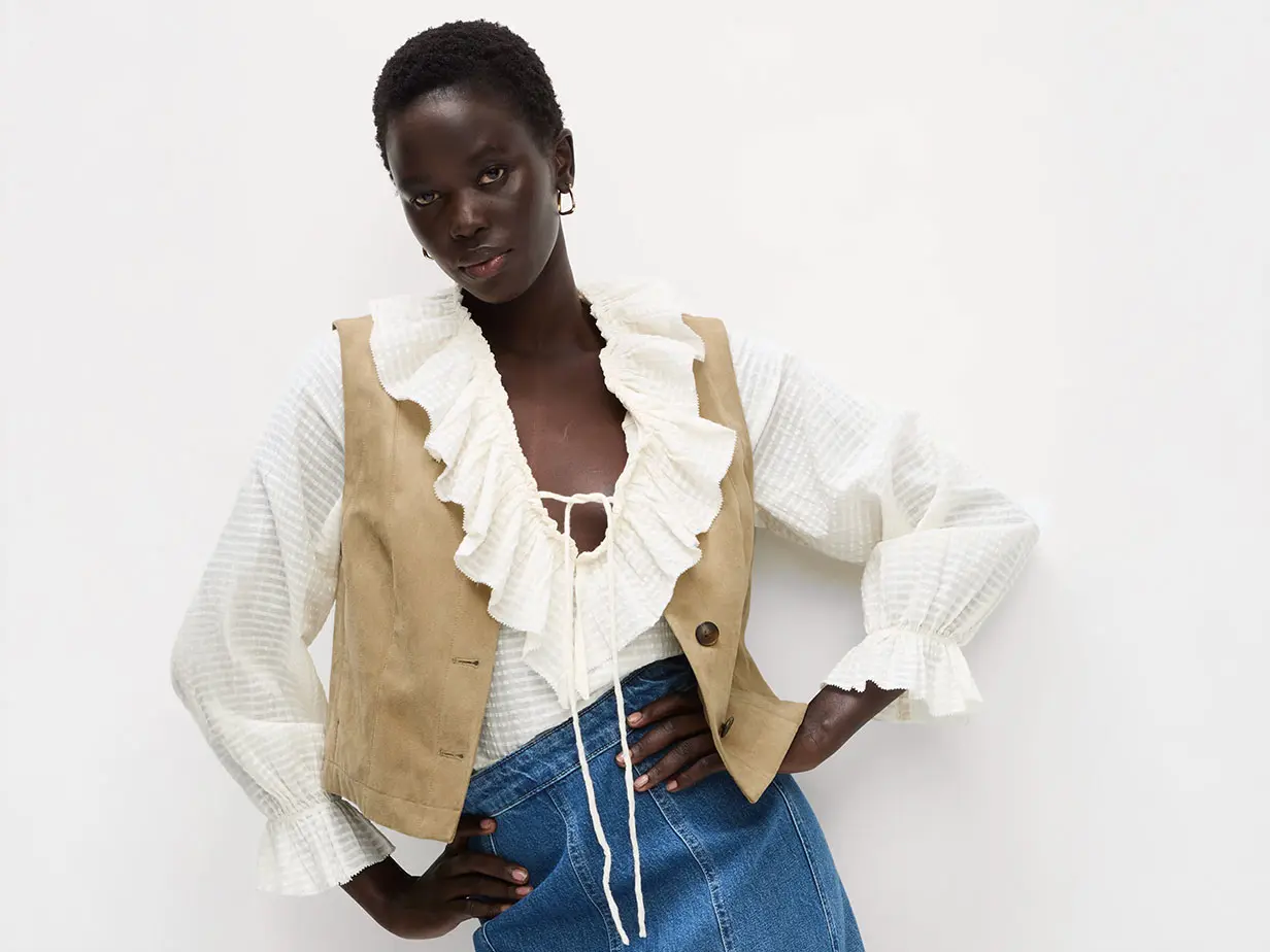 Woman wearing a ruffled blouse with a suede gilet, denim skirt and gold hoop earrings.