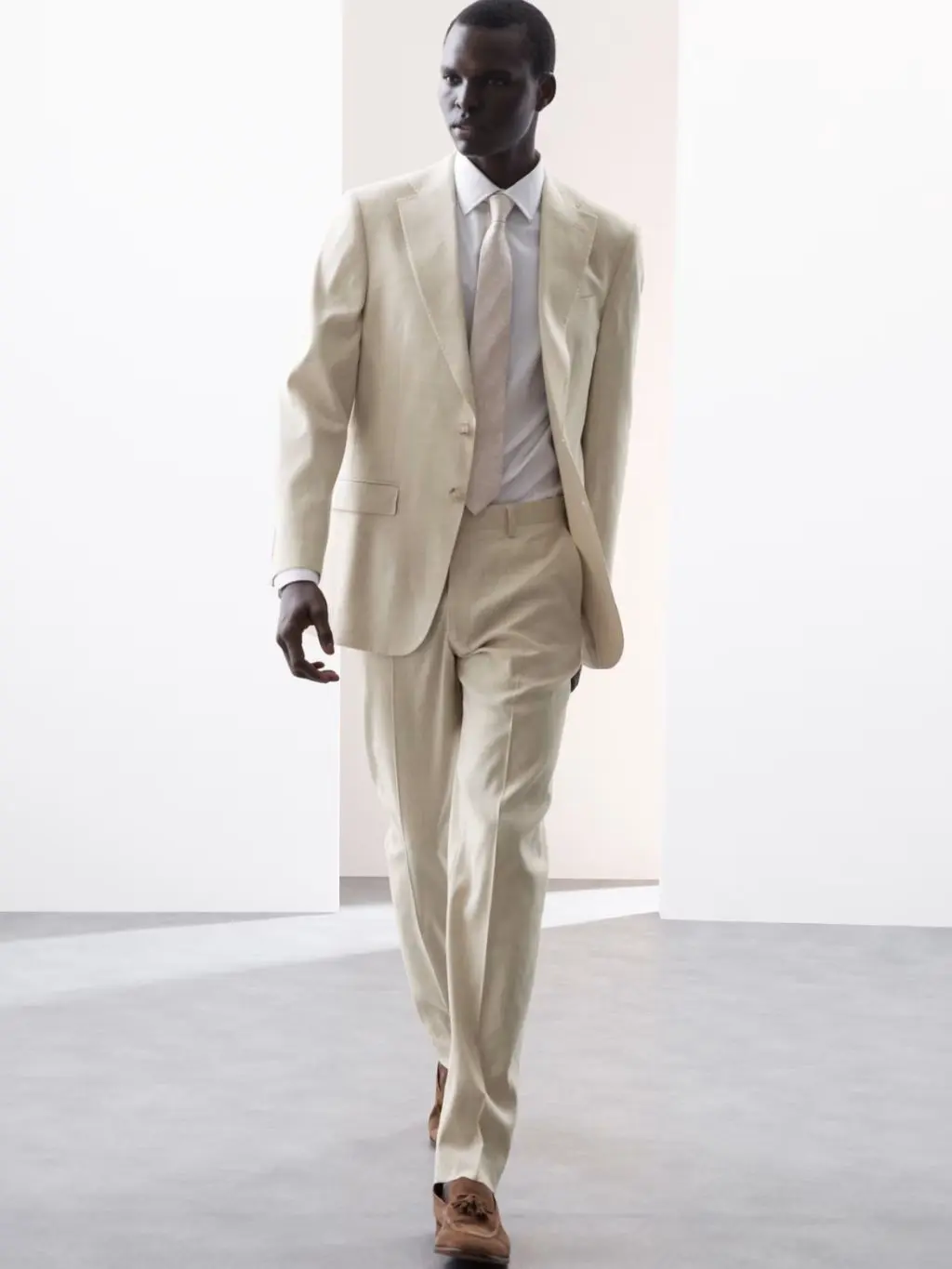 A man wears a pale cream linen suit and tie with brown shoes