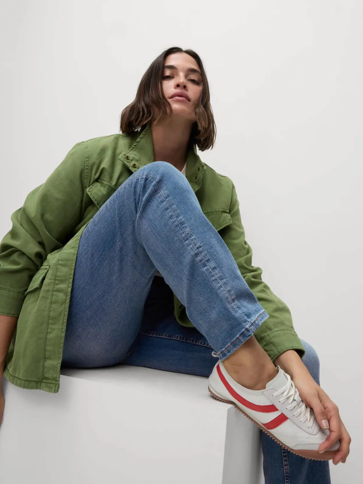 Woman sitting down, wearing green jacket, blue jeans and white trainers with red stripe detail 