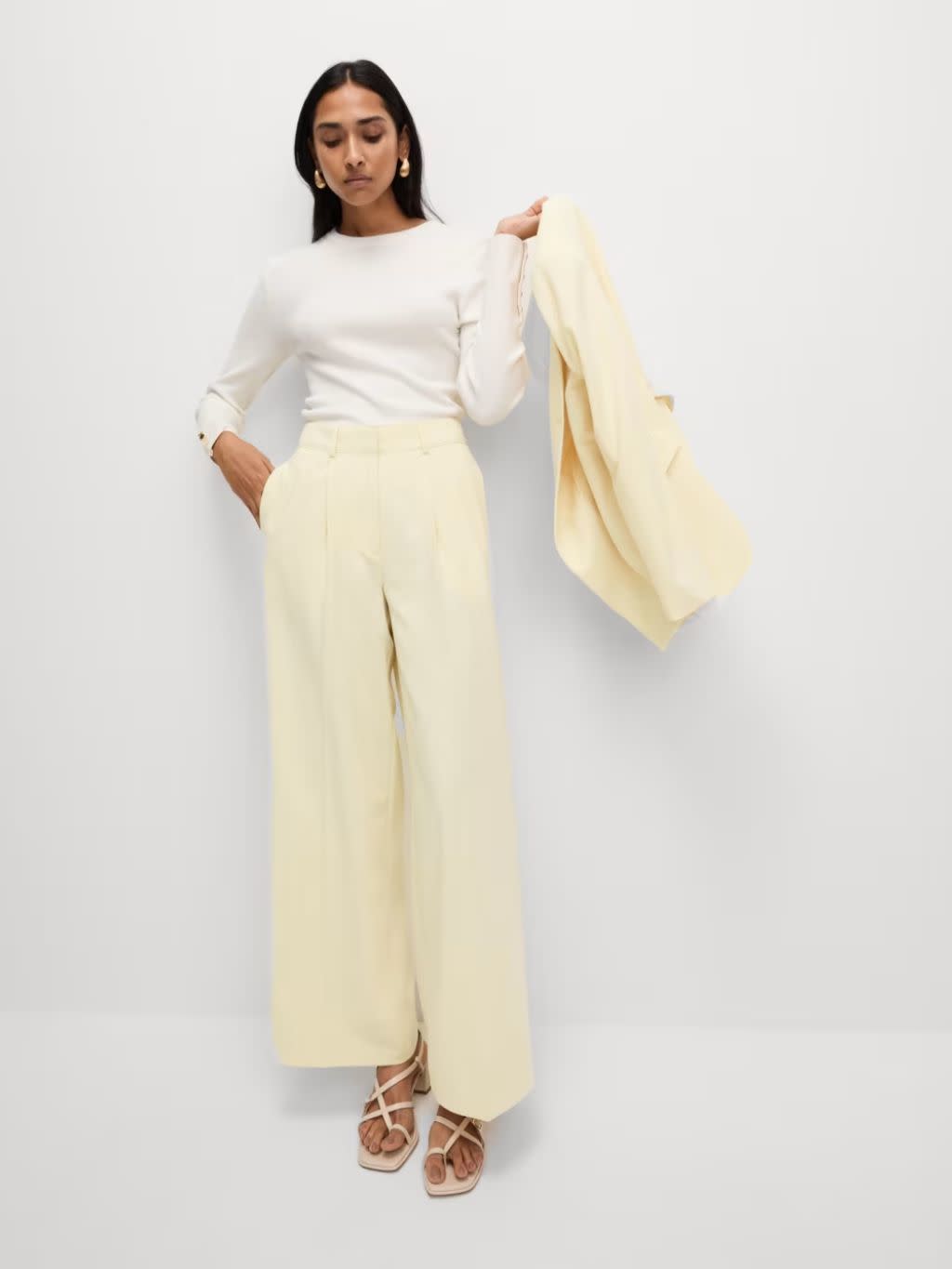A woman wears a cream top with wide leg lemon trousers and a matching jacket over her shoulder