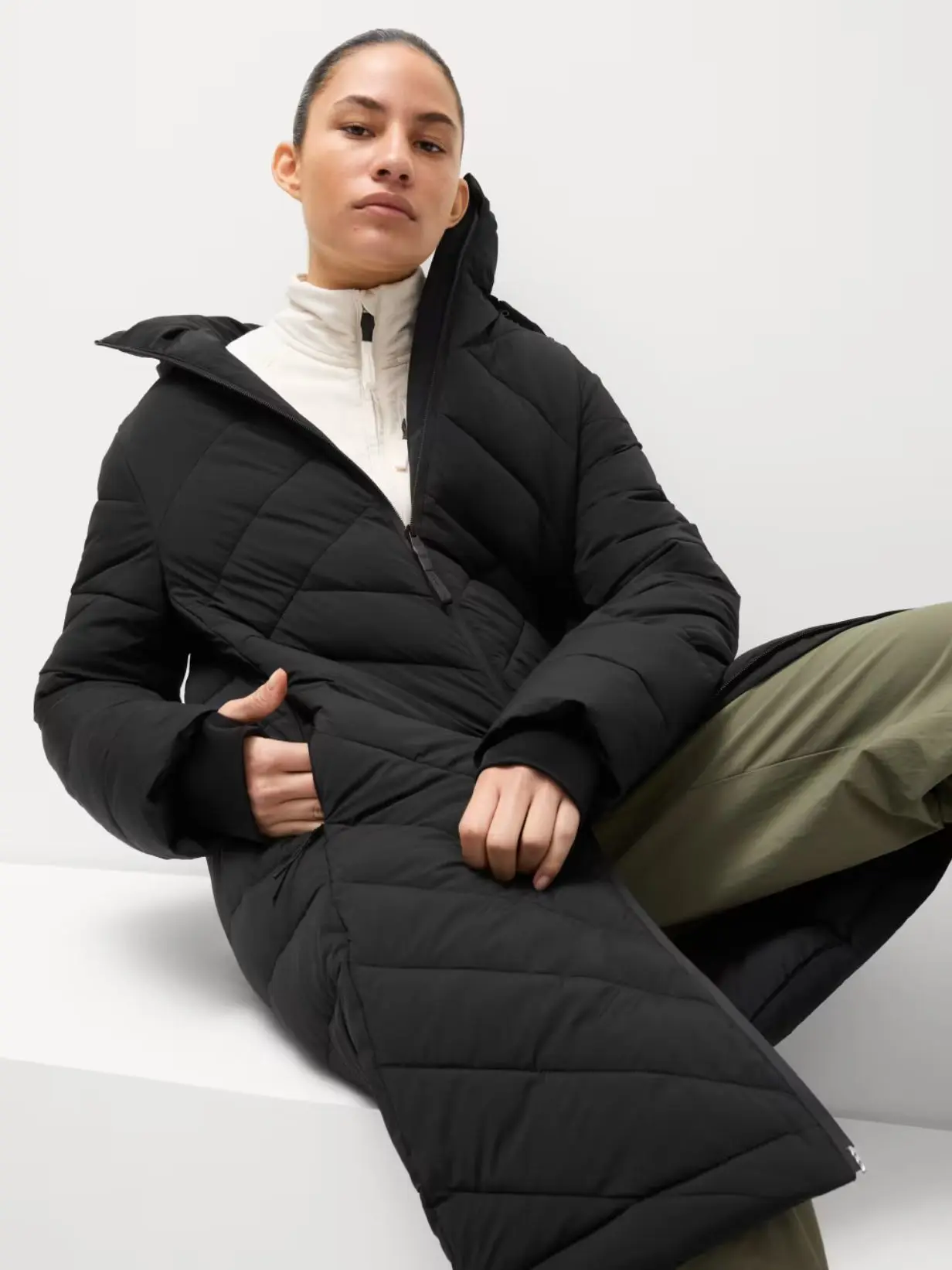 A model sat down wearing a black long puffer coat with a white top and khaki trousers