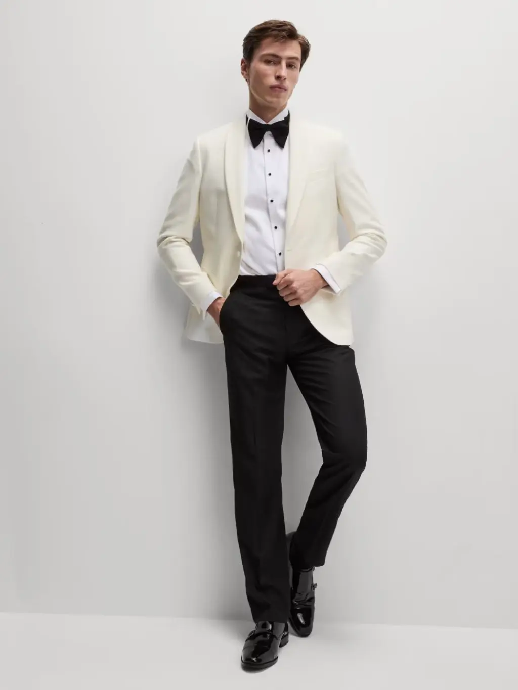 A man is wearing a white tuxedo jacket, black bow tie, white shirt and black trousers stood against a grey wall