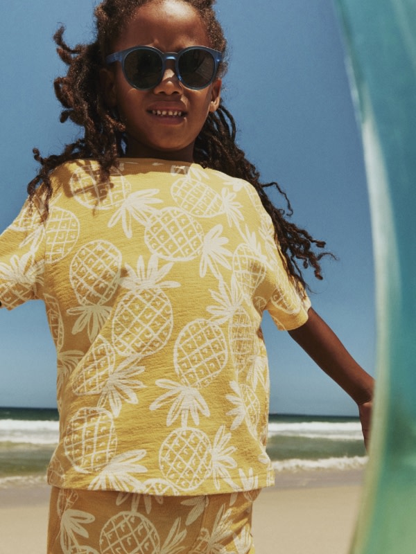Boy wearing a yellow and white pineapple print t-shirt and matching shorts. Shop boys’ tops