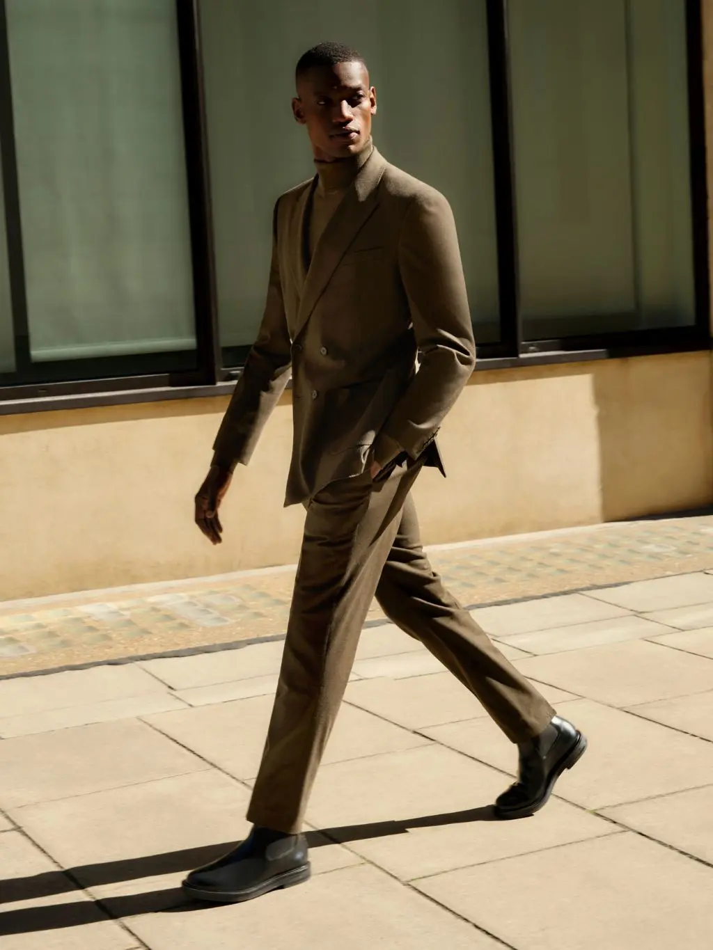 A man walks on a street in front a building wearing a brown suit and chelsea boots