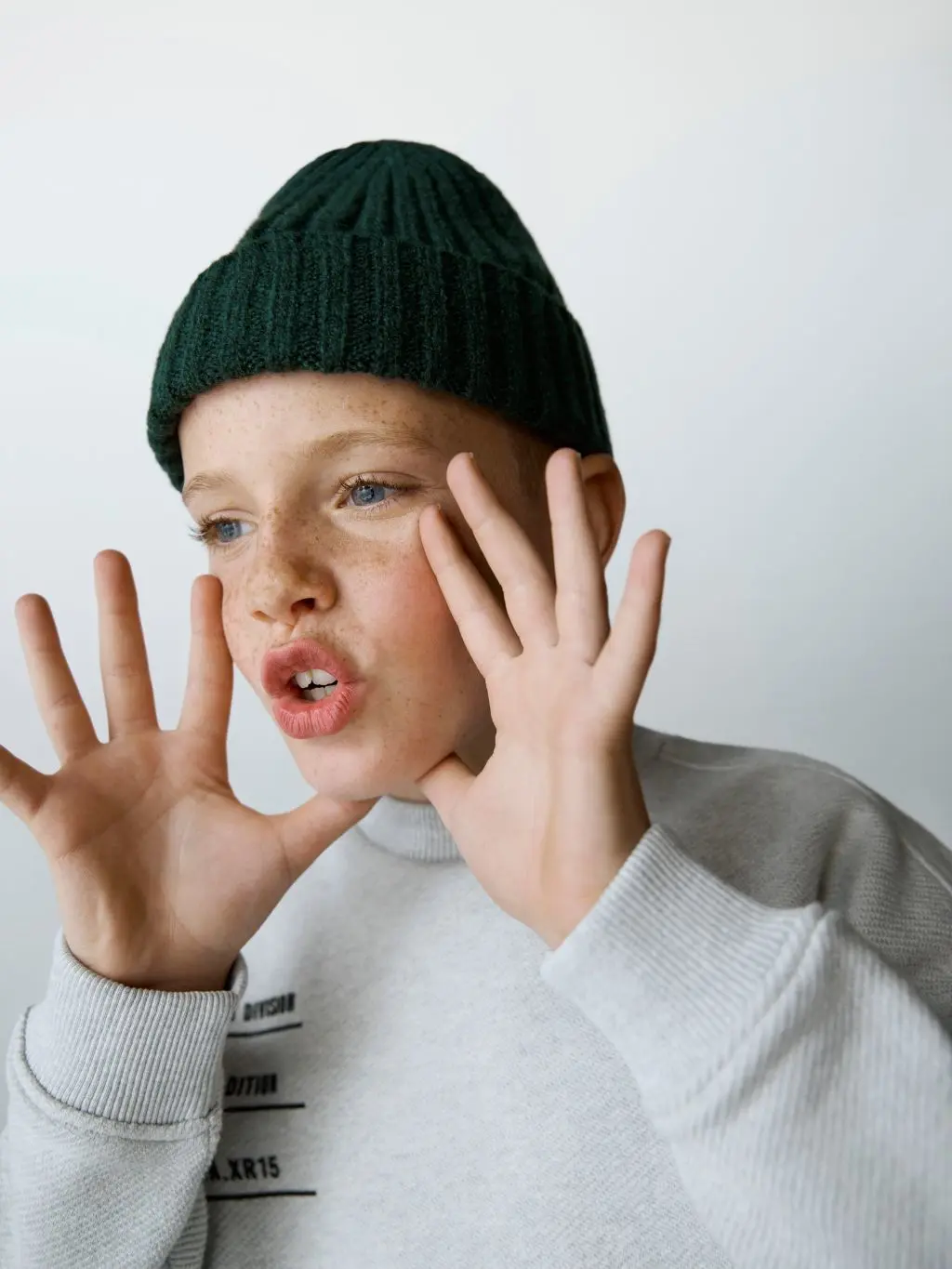 a boy shouts to one side while wearing a dark green beanie and grey sweatshirt