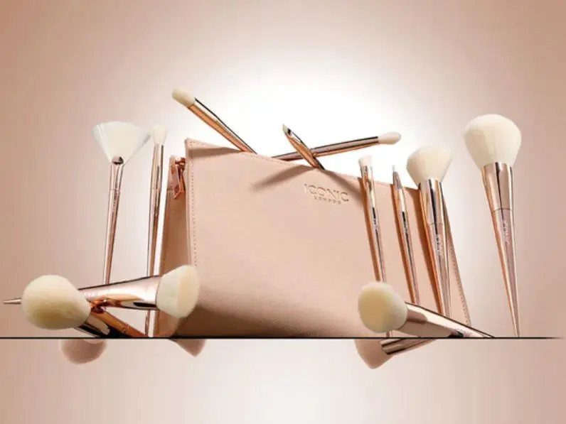 A product shot of rose gold and cream makeup brushes, with a makeup bag, set against a pink backdrop