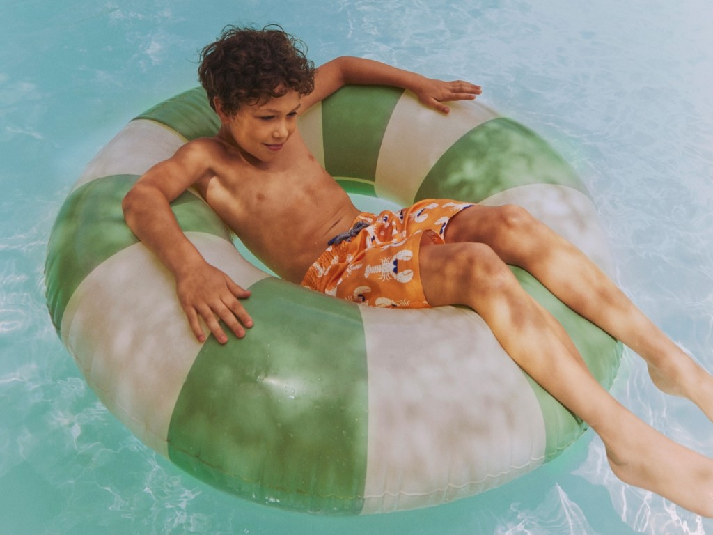 Boy wearing orange patterned swimming trunks lying in a green and white striped rubber ring. Shop boys’ swimwear