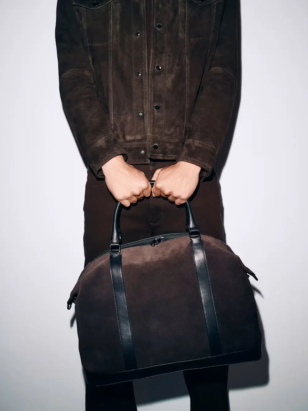 A man standing in a a brown suede trucker and brown trousers carrying a suede holdall bag