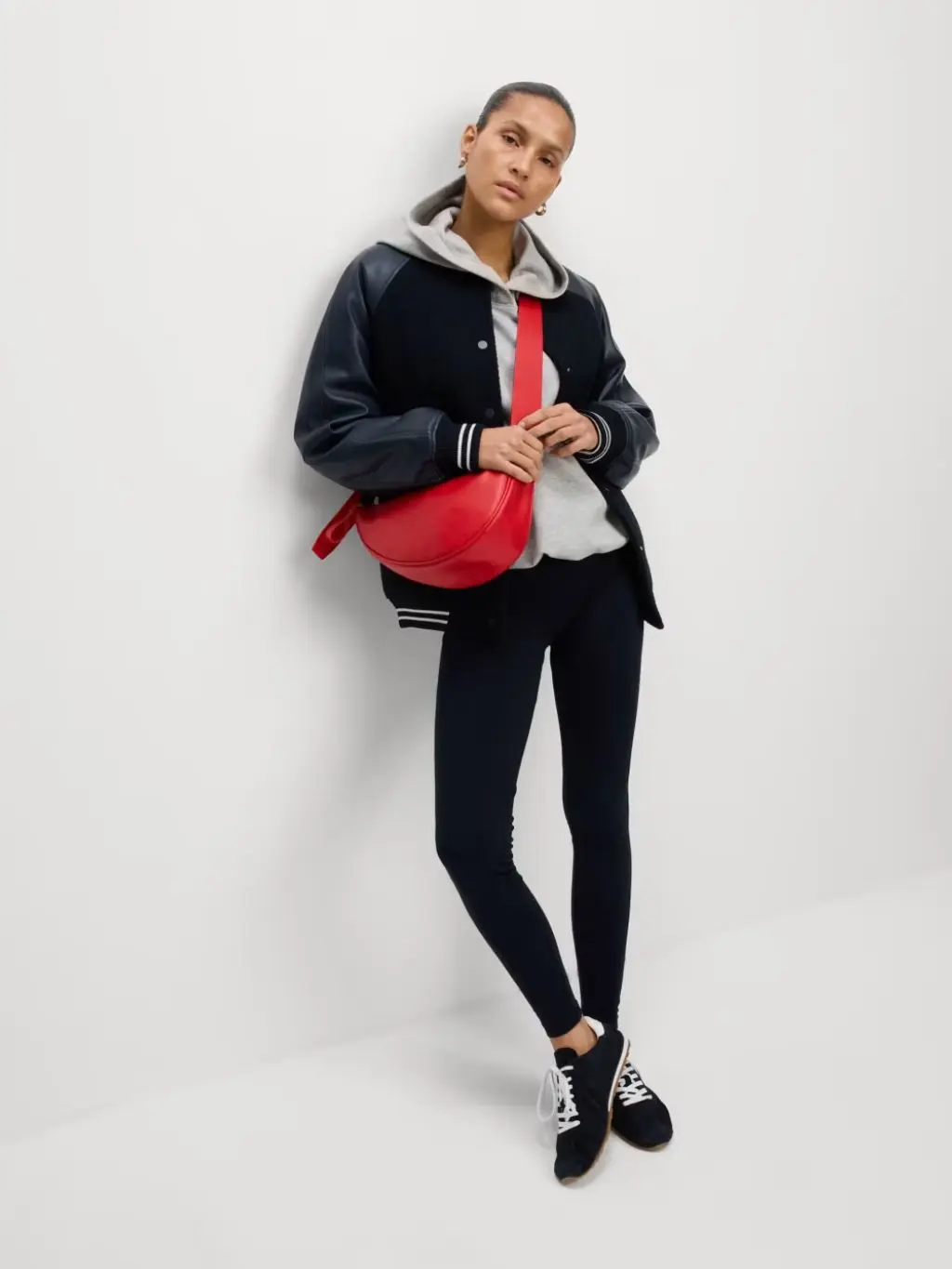A woman wears a baseball jacket over a hooded sweatshirt and leggings along with a red crossbody bag