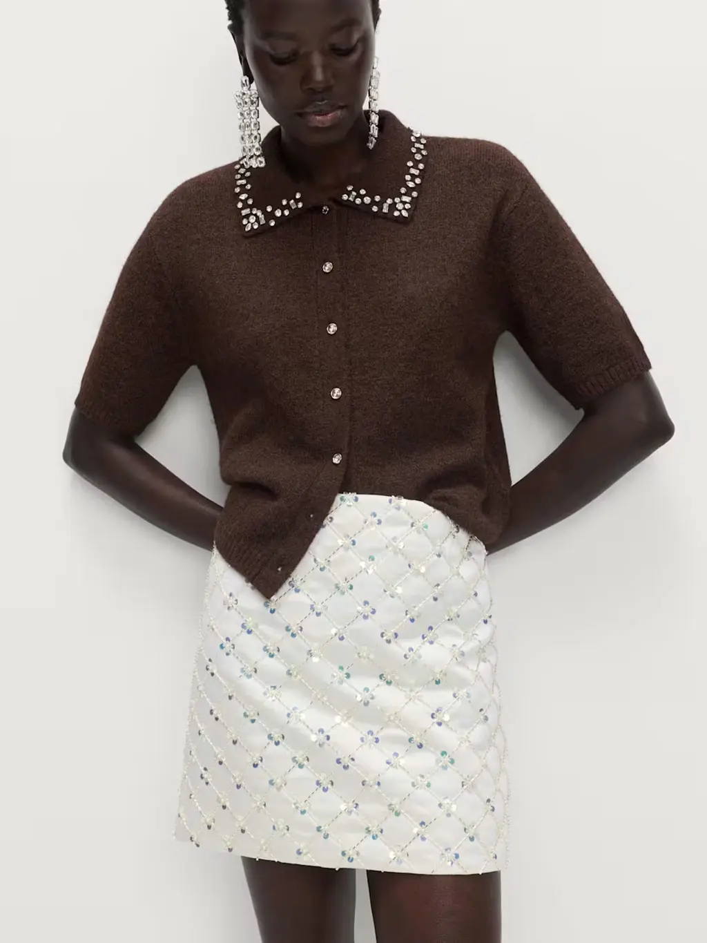 Woman wearing an embellished white satin skirt with a sequinned brown cardigan and drop earrings