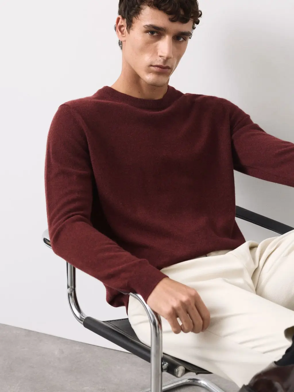 A man sat on a chair wearing a dark red cashmere jumper and white jeans