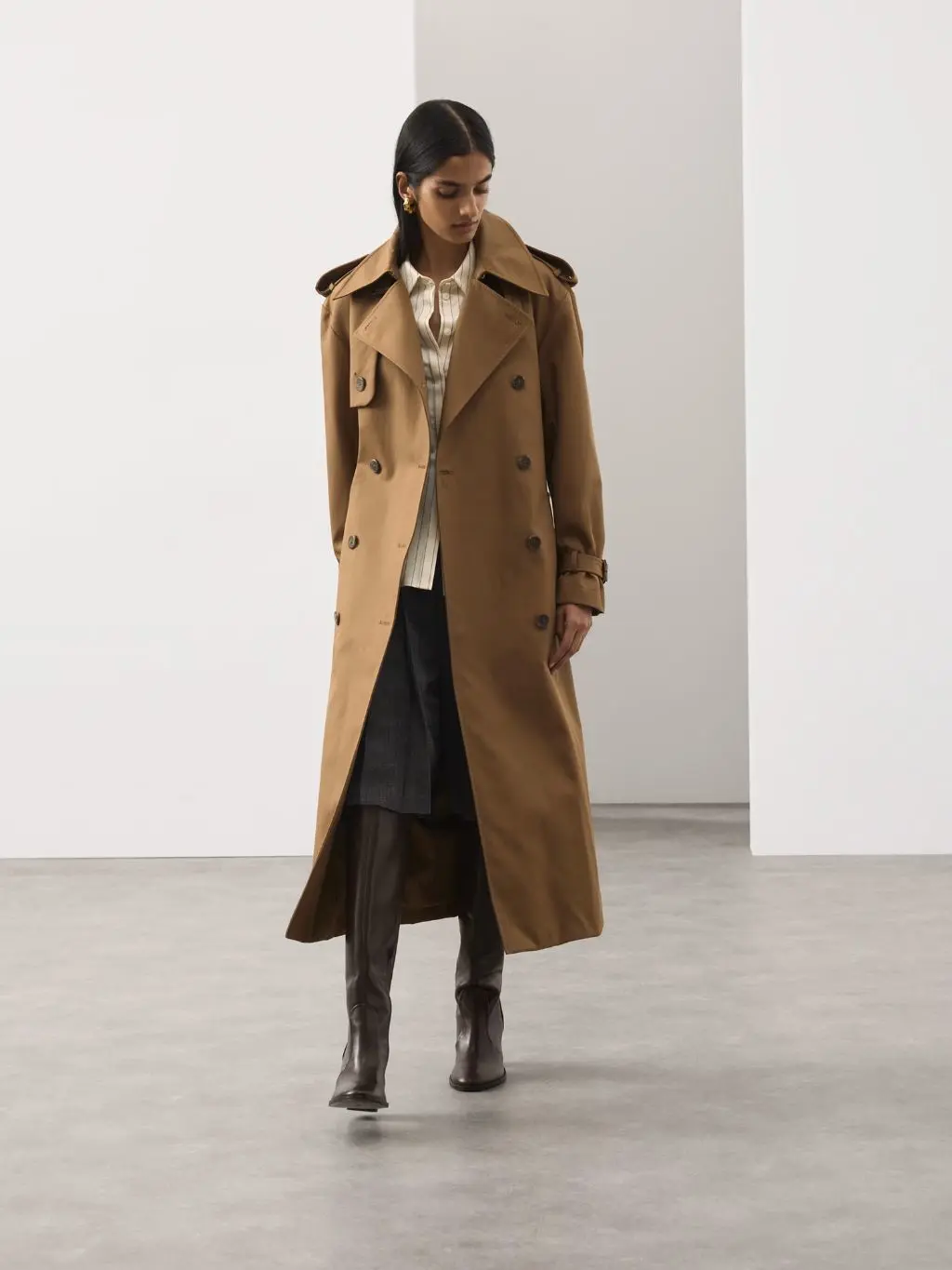 A woman wears a tan trench coat over a blouse and skirt, she is looking down towards the floor
