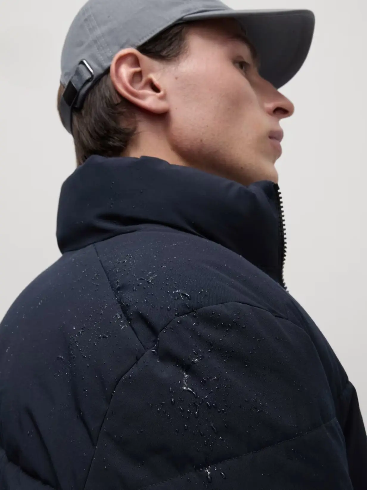 A man wearing a grey baseball cap and navy blue puffer jacket with water on it