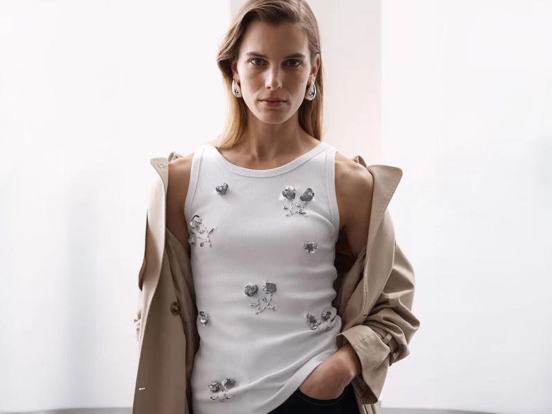 Woman wearing white sequinned vest top, silver dome earrings and a stone-coloured trench coat