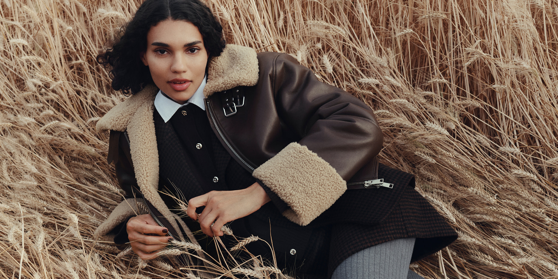 Woman wearing a brown aviator jacket and black dress in a field of wheat