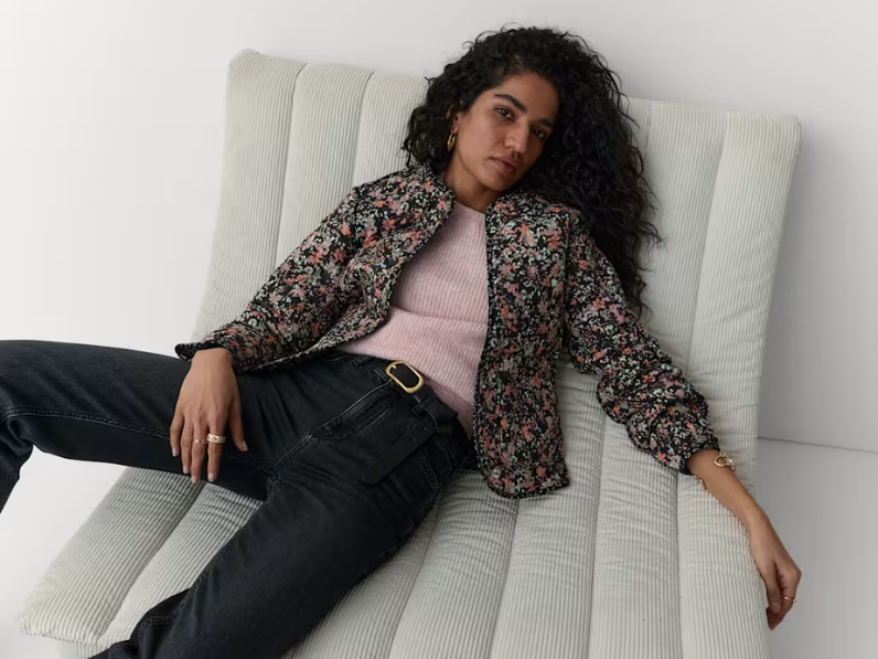 A woman sits on a chair wearing a floral quilted jacket by Per Una, pink jumper and jeans