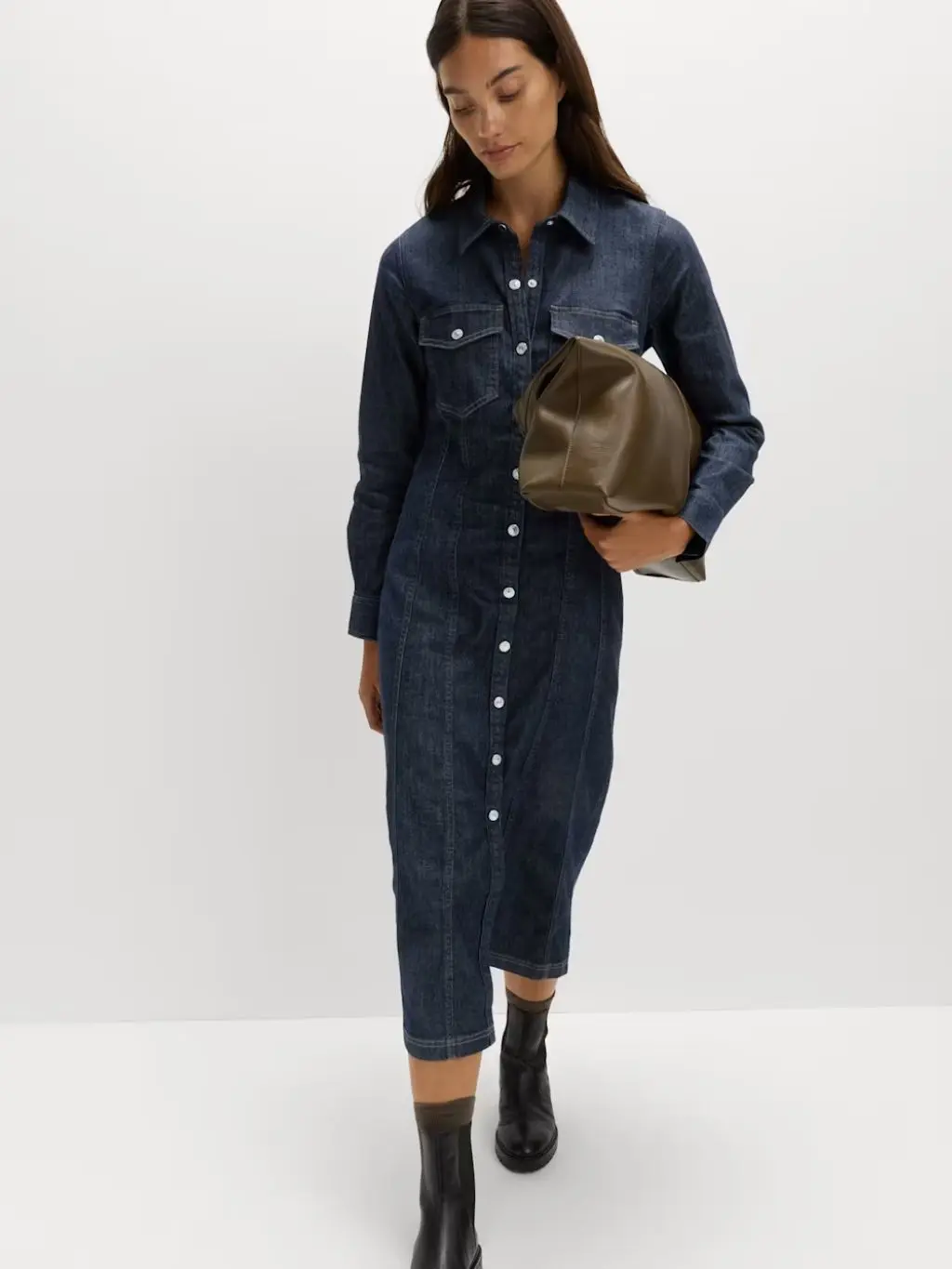 A woman wears a dark denim shirt dress and ankle boots, carrying a bag under her arm