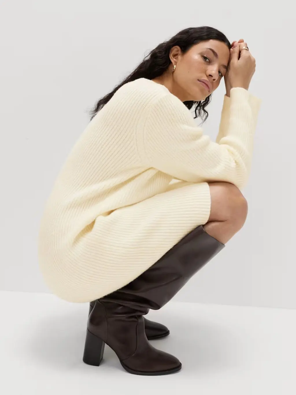 A woman wears a cream jumper dress with brown knee high boots and no tights