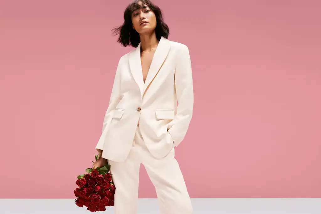 Woman wearing white suit holding a bouquet of red roses against a pink background
