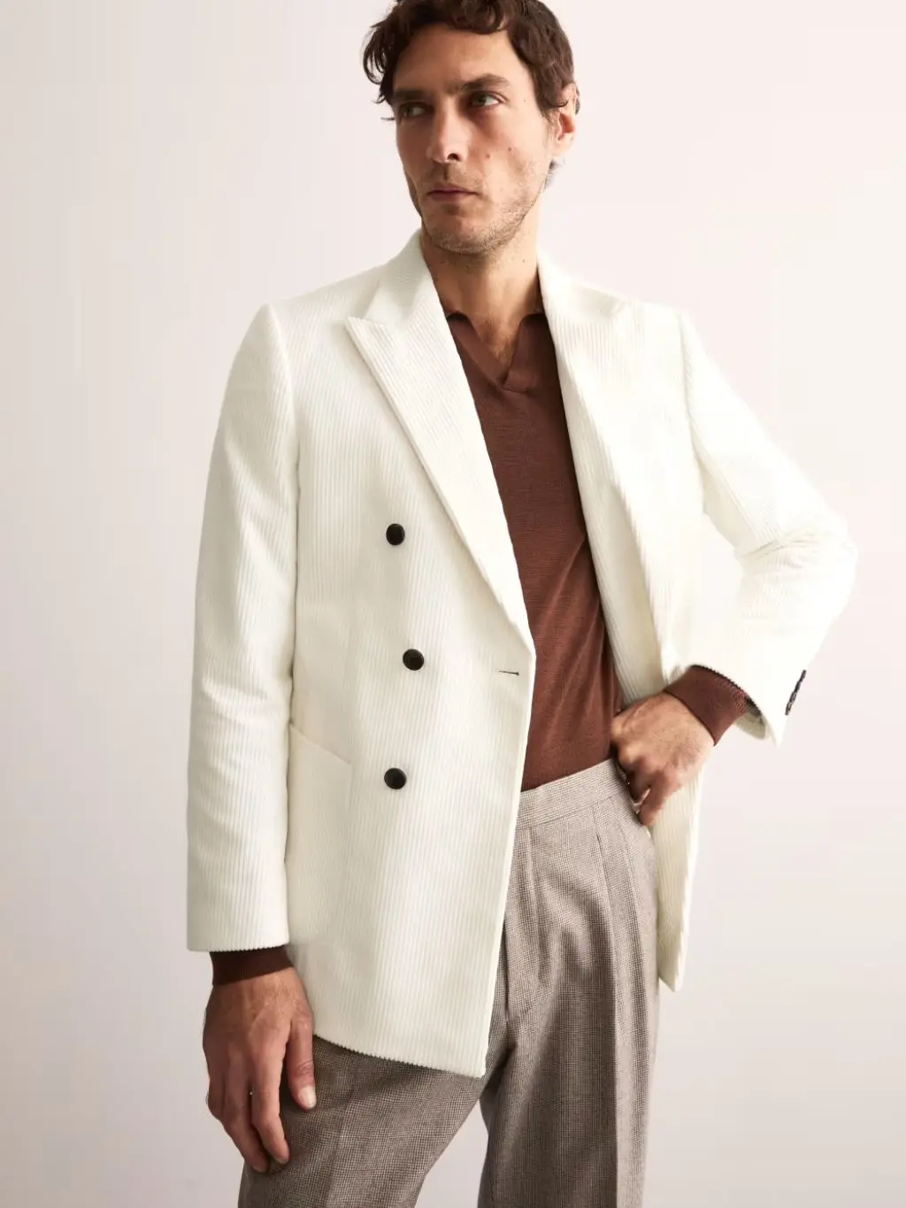 A dark haired model wearing a white cord blazer over a brown top and check trousers