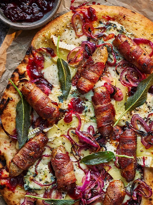 Close up of pigs and blankets on pizza with sage leaves and red onion