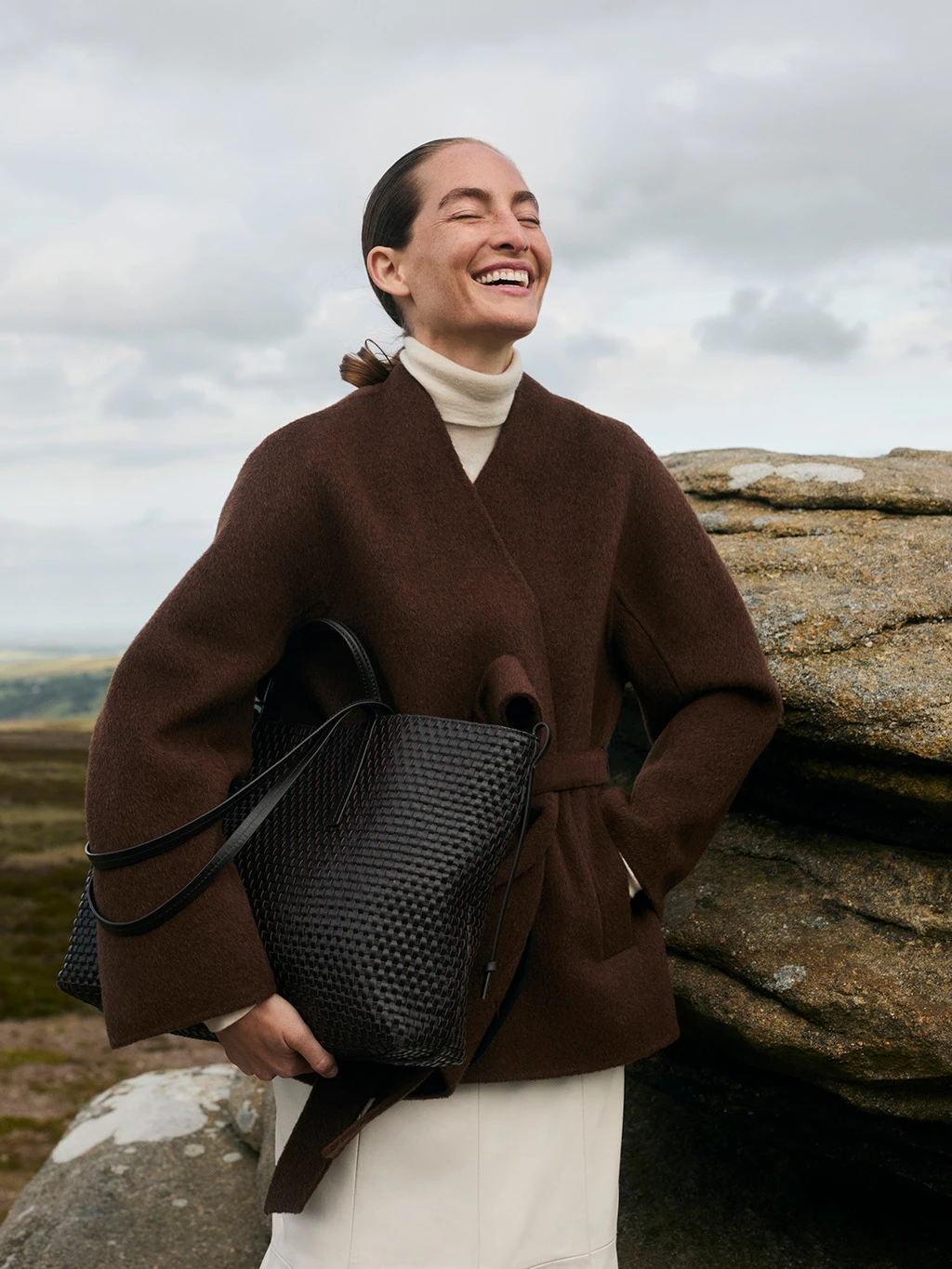 Woman wearing a brown blazer, cream polo neck and white skirt, holding a woven leather bag.