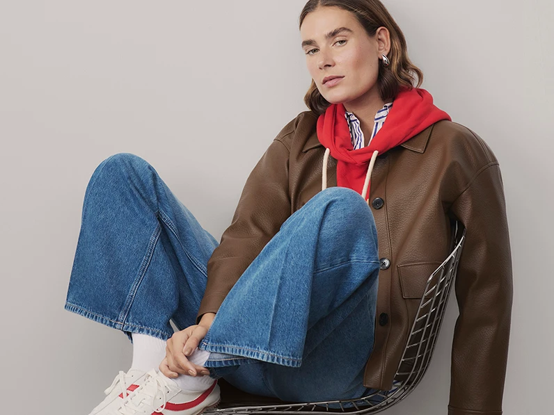 Woman wearing brown jacket, red hoodie, blue jeans and retro trainers.