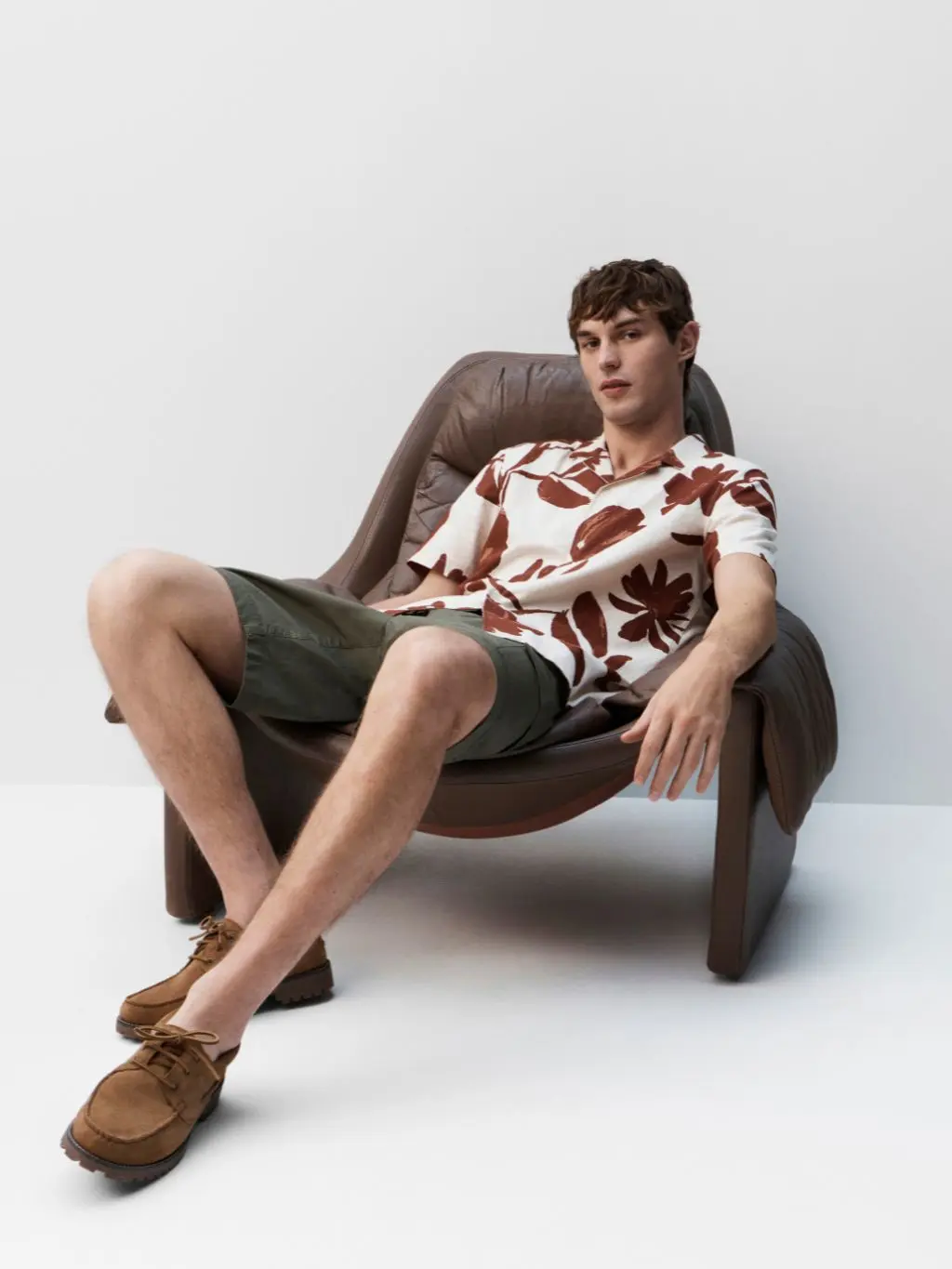 A man sits on a chair wearing a floral print short sleeve shirt, green shorts and suede loafers