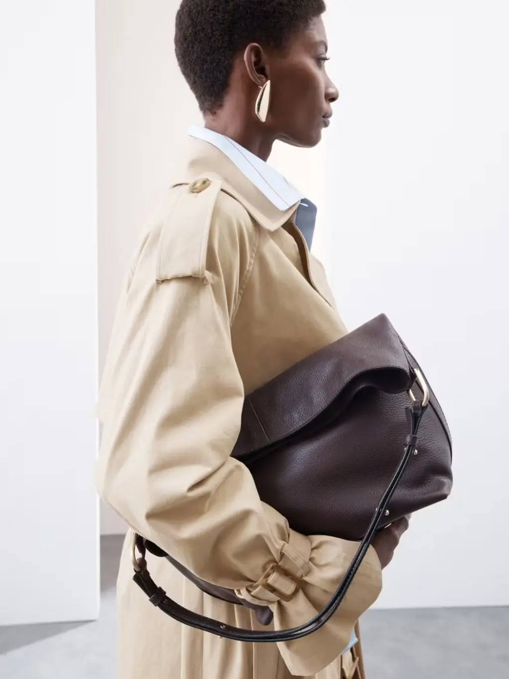 A woman carries a large bag under her arm, while wearing a trench coat over a blue shirt