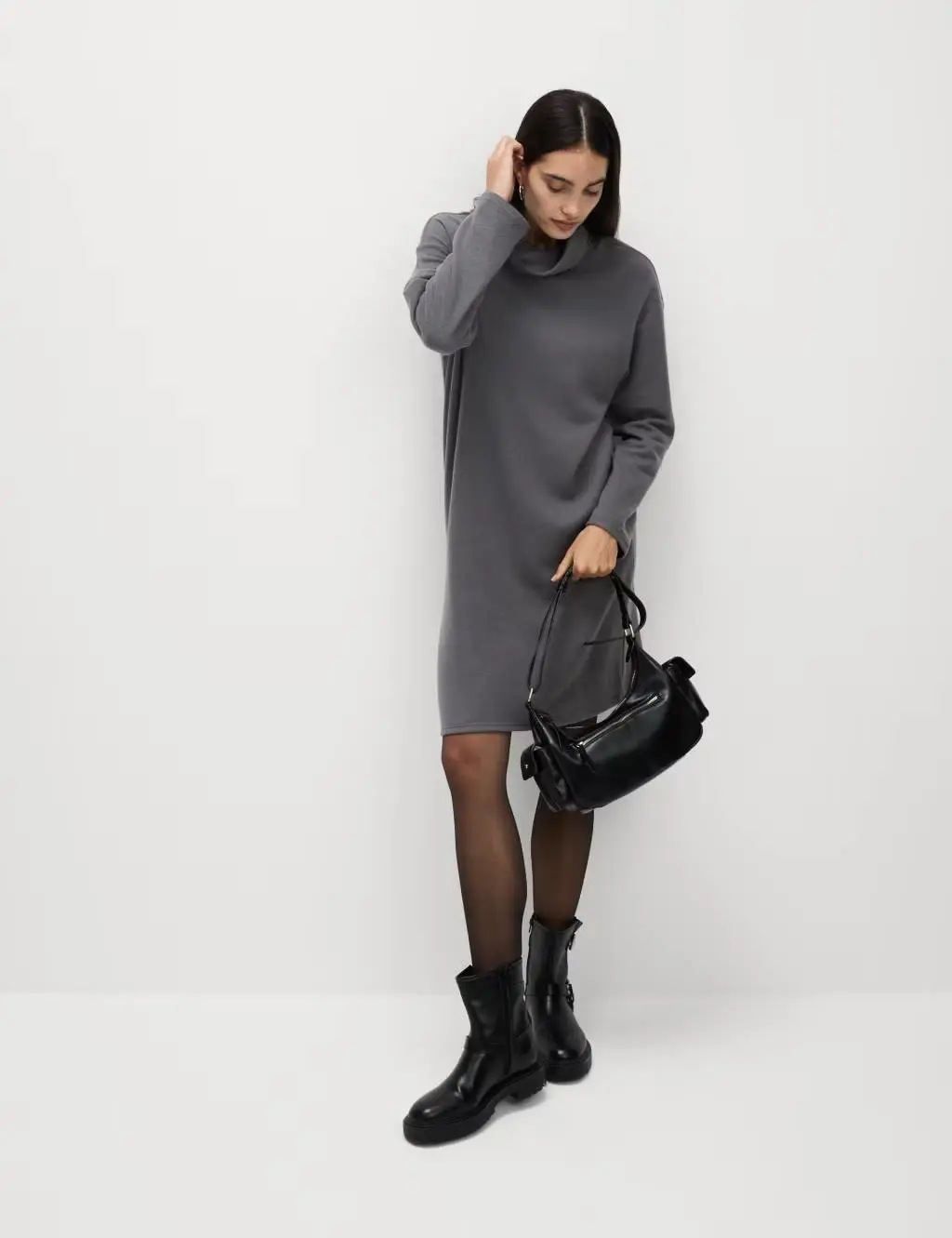 A woman wearing a grey knitted roll neck jumper dress, carrying a black handbag