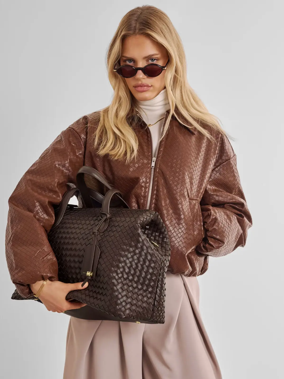 Woman wearing neutral outfit including smart trousers, a roll-neck top and jacket, holding a large brown woven bag