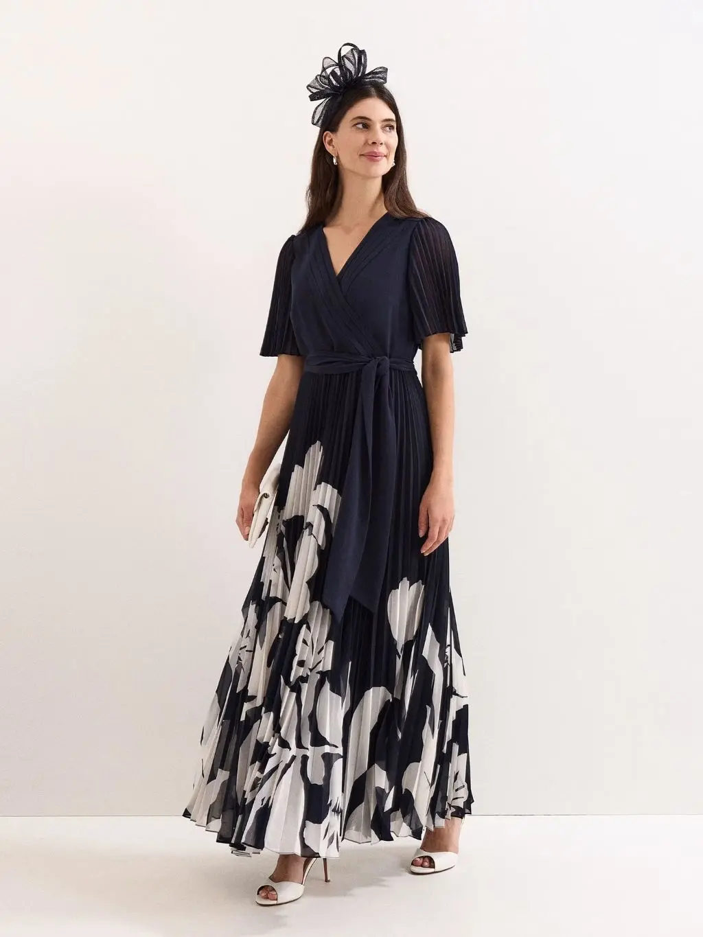 A woman wears a navy and white floral wrap dress with a fascinator and heels