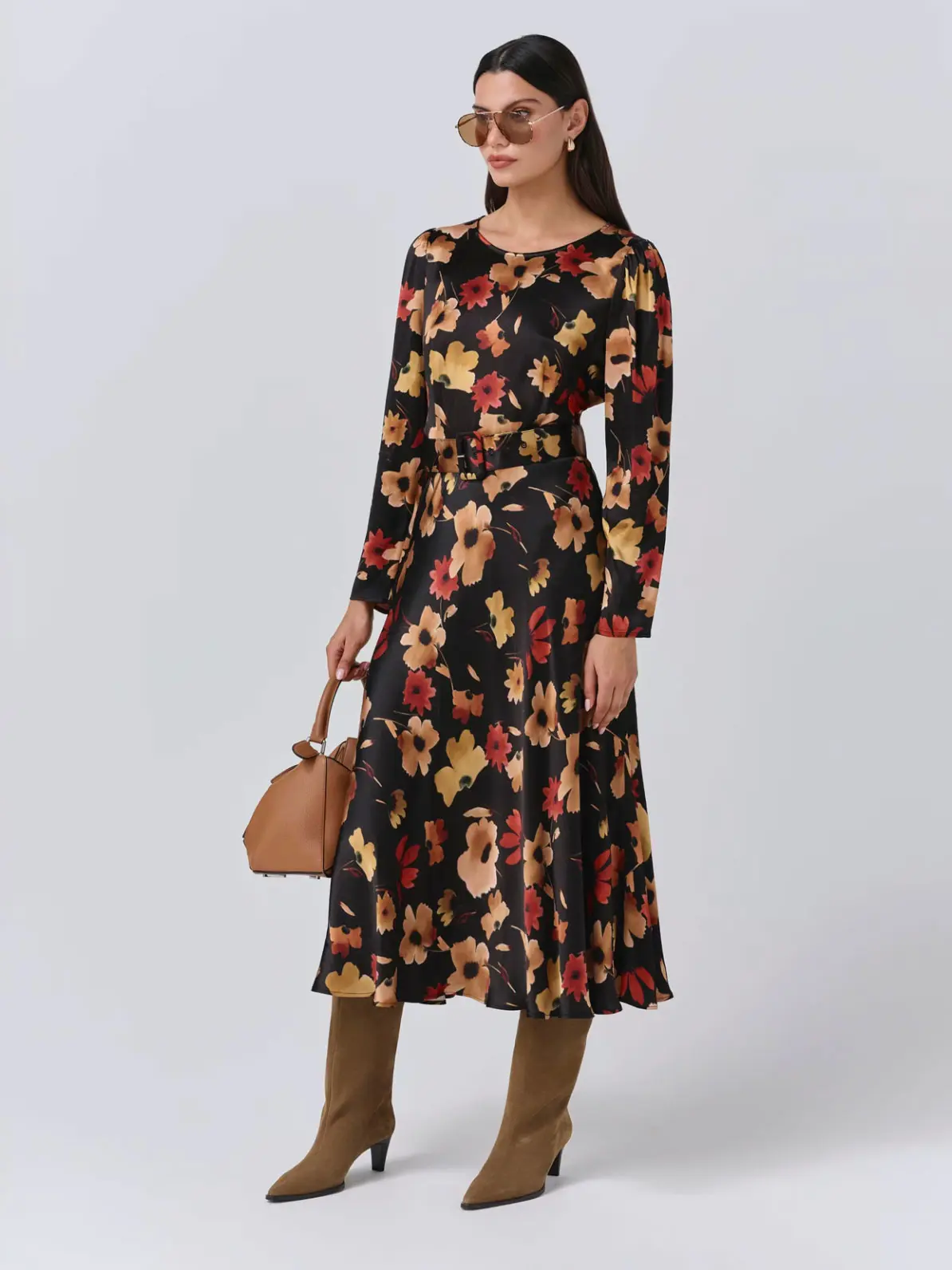 Woman wearing black, gold and red floral dress, brown heeled knee-high boots, holding brown bag and wearing sunglasses