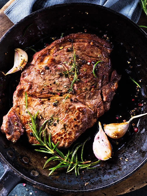 A pan with a cooked piece of steak, garlic cloves and rosemary.
