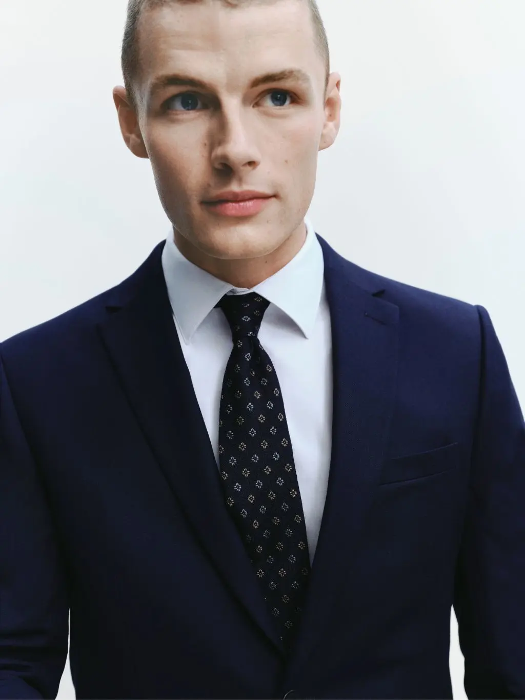A man with a tie, white shirt and blue suit on looks away from the camera