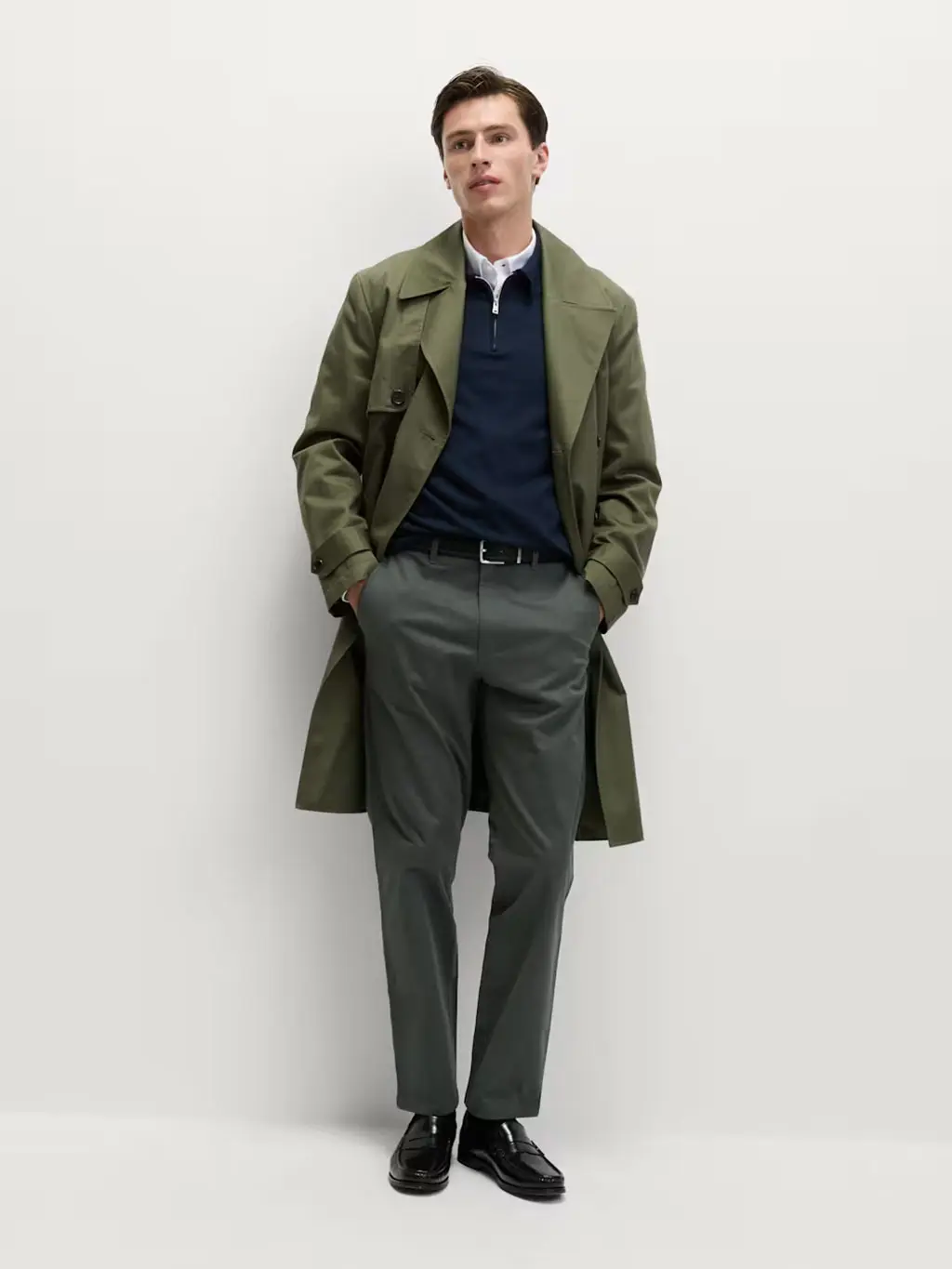 Man wearing grey chinos with a white shirt, quarter-zip jumper, trench coat, black belt and loafers