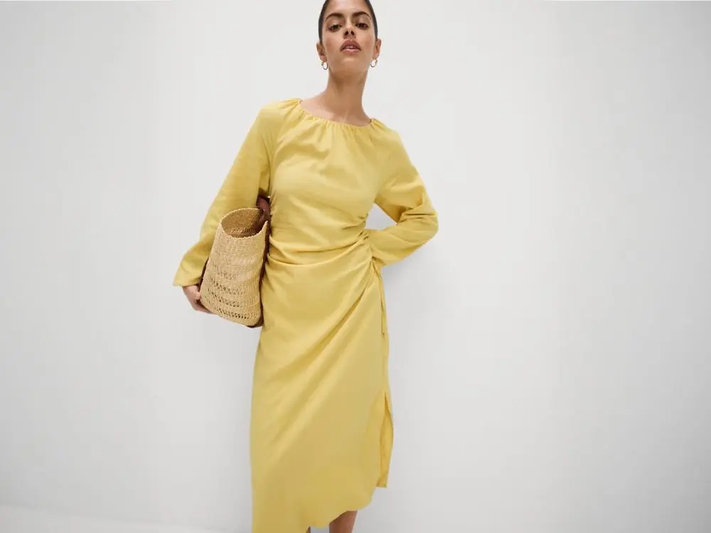 A woman weras a butter yellow linen dress, carrying a straw tote under her arm
