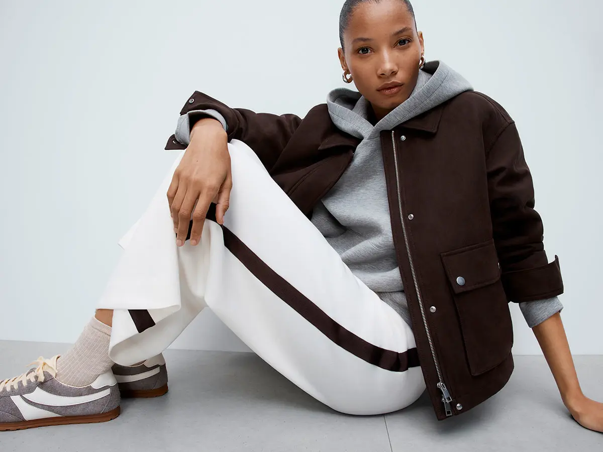 Woman wearing grey hoodie under brown jacket, white trousers with brown stripe detail and light neutral trainers 