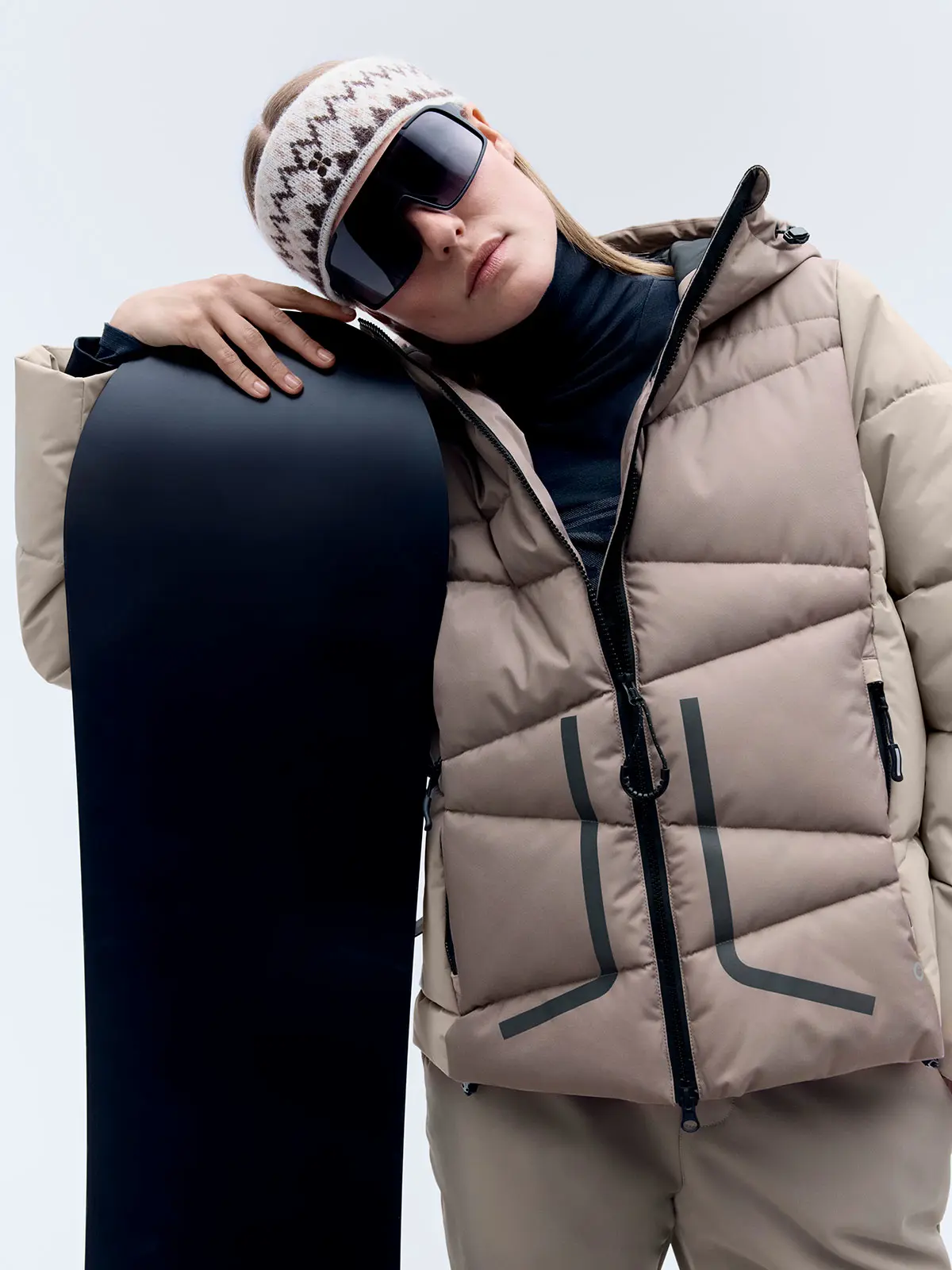 Woman wearing neutral ski jacket, neutral trousers, patterned headband and ski glasses, holding black snowboard