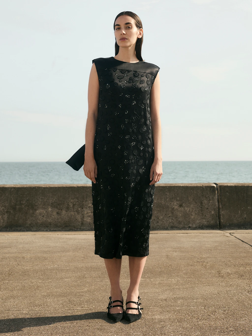 A woman wears a black Jaeger evening dress while standing on a beach