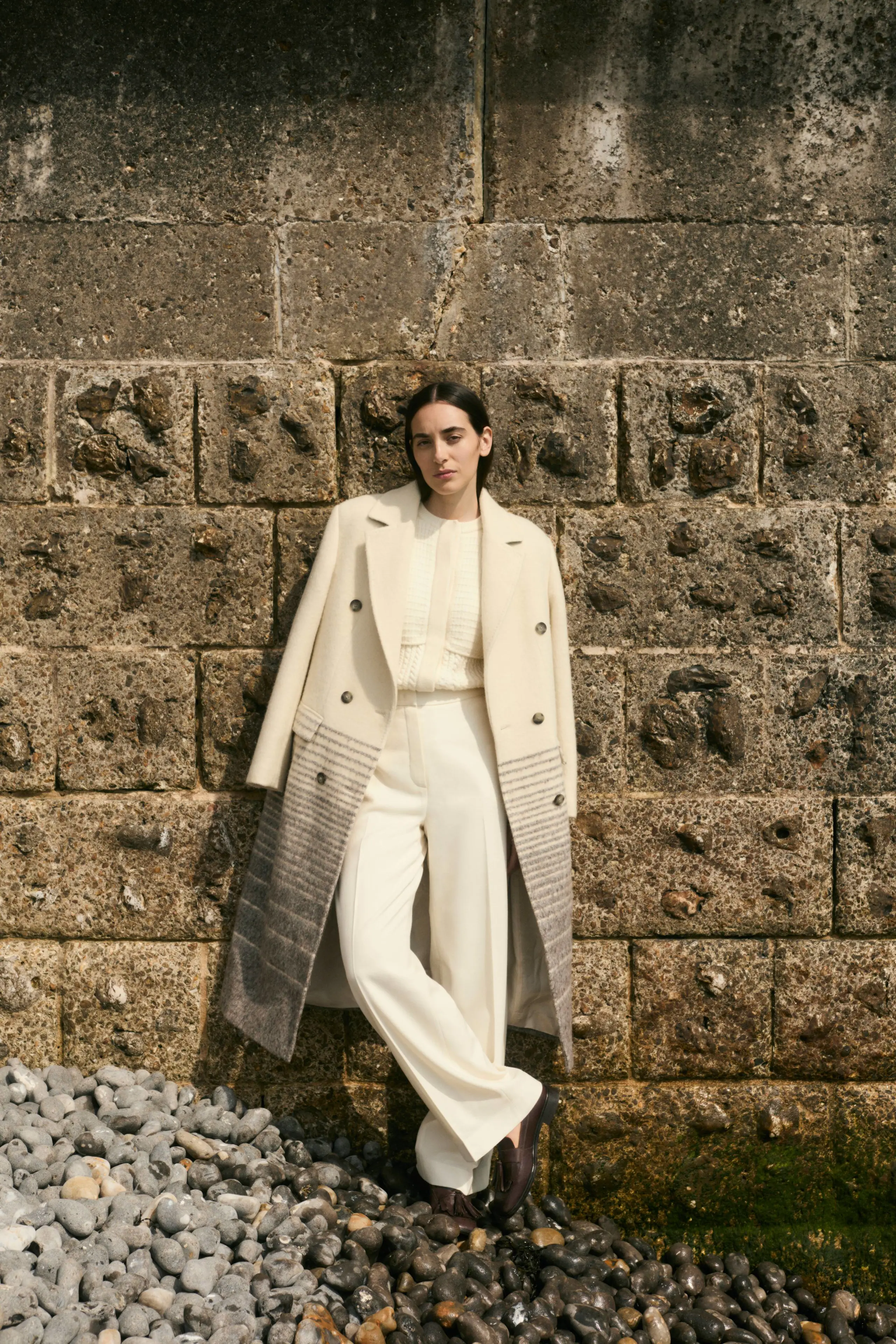 A woman wears a cream coat, cardigan and trousers while standing on a beach against a sea wall
